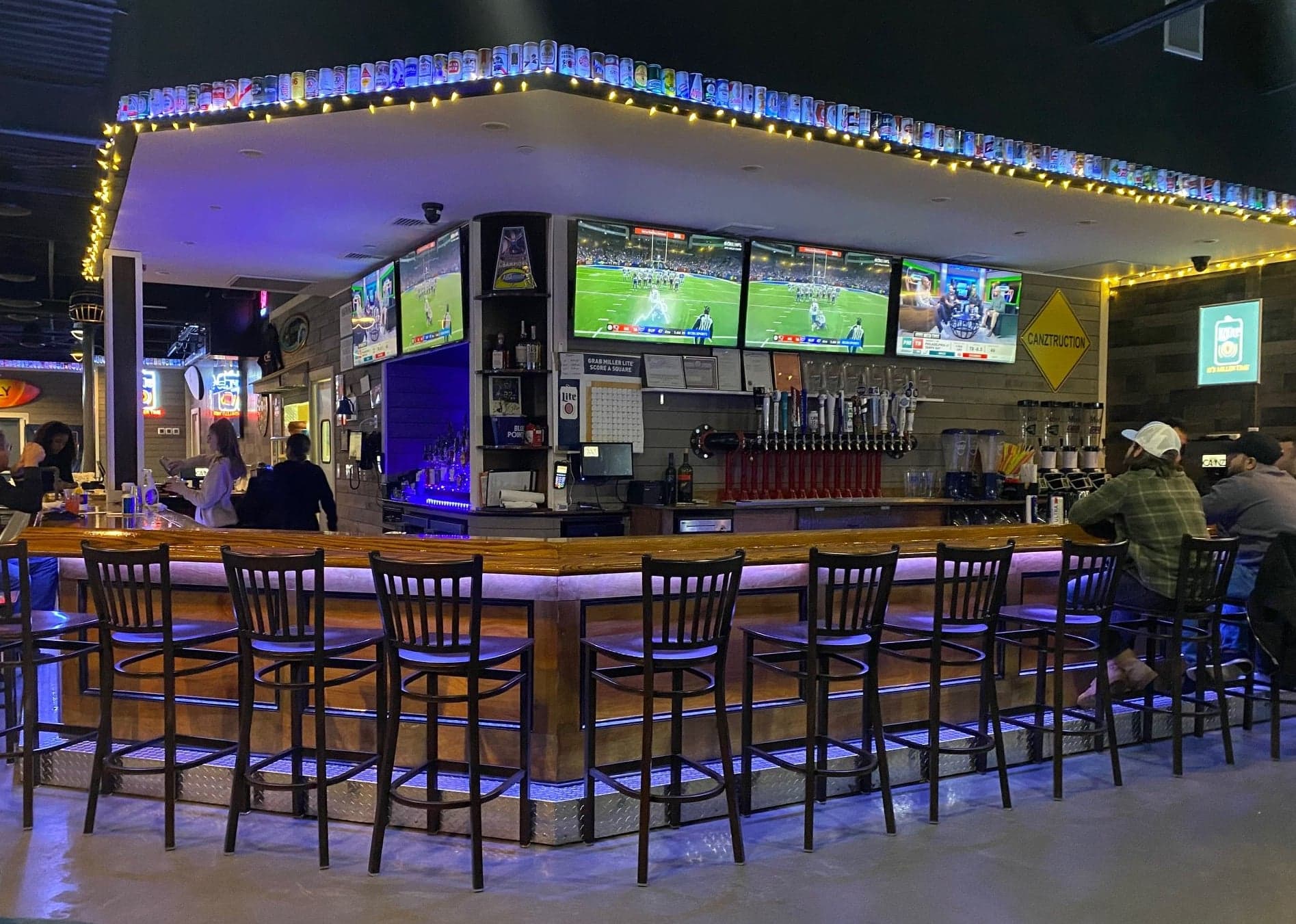 Wraparound bar at Canz Bohemia with blue-lit stools, football on wall-mounted TVs, a row of beer taps and a shelf of beer cans lined with string lights.