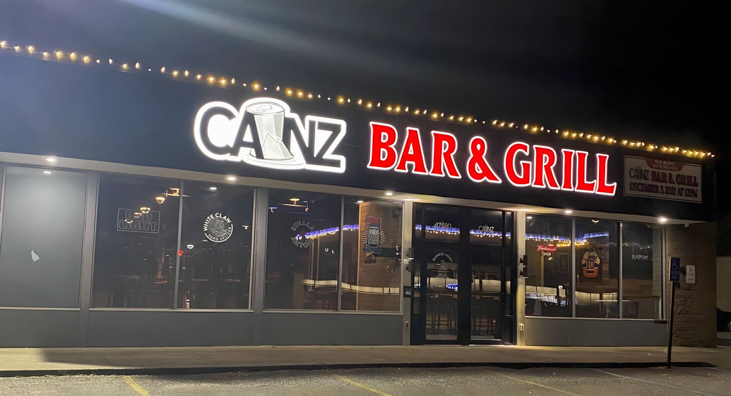 Canz Bar & Grill storefront at night, its illuminated sign glowing above string lights and big front windows.